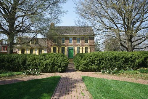 A historic yellow brick house surrounded by green hedges and trees, with a brick pathway leading to the entrance.