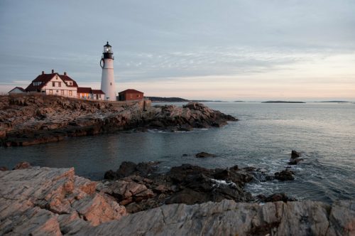 A lighthouse stands on rocky shores at sunset, with calm waters and a cloudy sky in the background.