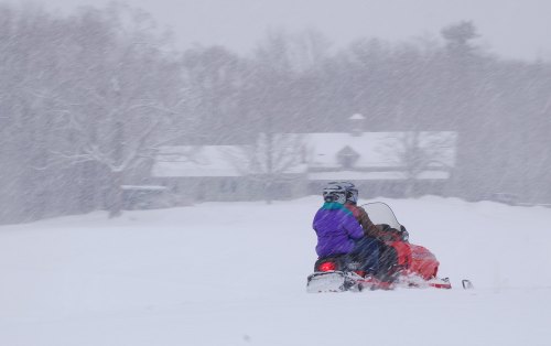 Two people ride a snowmobile through heavy snowfall, with a house visible in the background.
