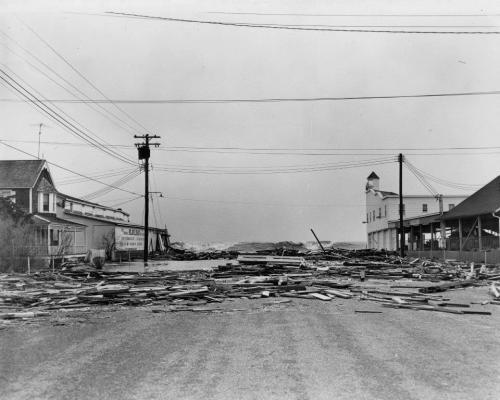 A desolate street lined with debris and damaged buildings, with power lines overhead in a black-and-white photo.