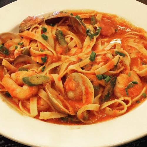 A bowl of fettuccine pasta with shrimp, clams, and green vegetables in a rich tomato sauce, garnished with herbs.