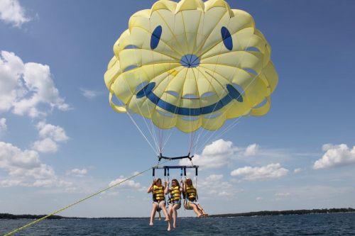 Four people in yellow life vests are parasailing under a large smiley face parachute over a lake.