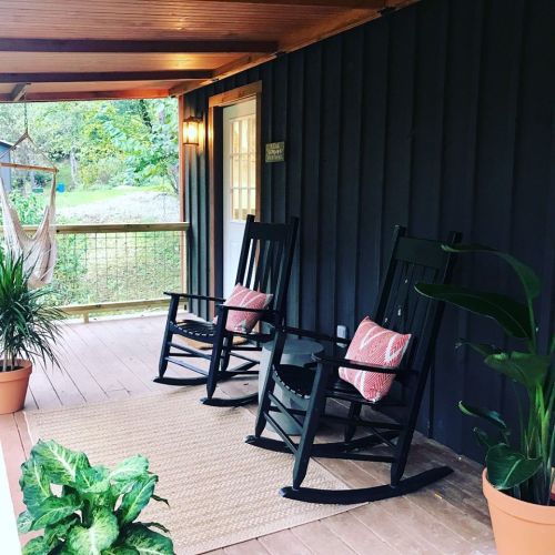 Two black rocking chairs with pillows on a porch, surrounded by potted plants and a cozy rug.