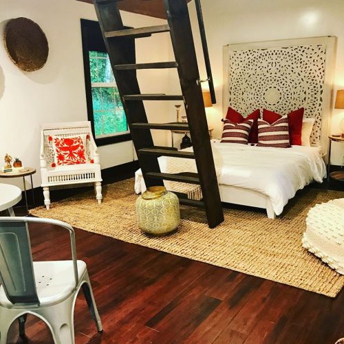 Cozy bedroom with a lofted bed, decorative pillows, a woven rug, and a small dining area. Warm, inviting decor.