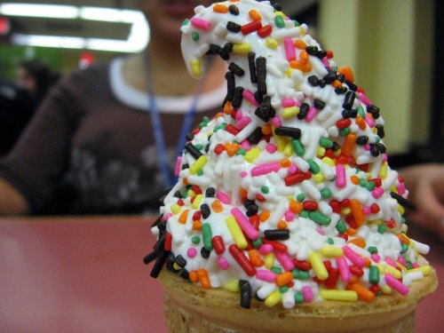 A close-up of a colorful ice cream cone topped with a swirl of whipped cream and a variety of sprinkles.