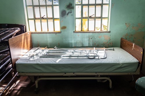 An empty hospital bed with a metal frame resting on a mattress, in a room with peeling paint and sunlight through windows.