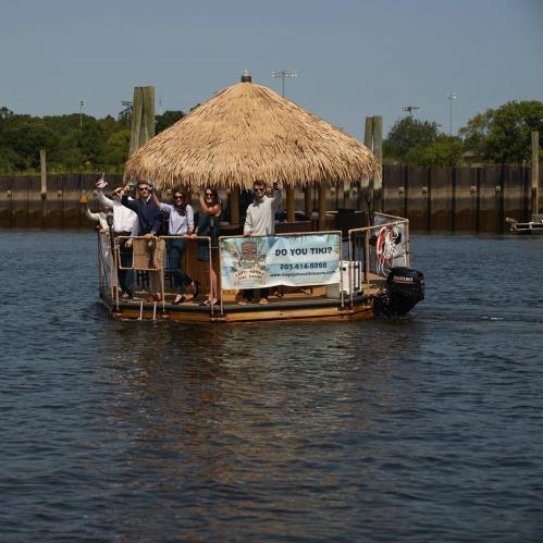 A group of people waves from a tiki boat on the water, with a thatched roof and a sign reading "Do You Tiki?"