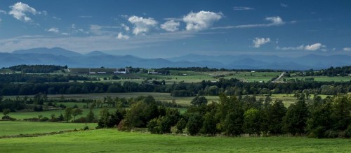 A panoramic view of rolling green fields and distant mountains under a blue sky with scattered clouds.