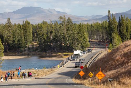 A scenic road with vehicles, surrounded by trees and mountains, near a lake with people observing the view.