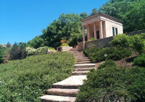 A stone pathway leads up to a small, classical-style building surrounded by lush greenery and trees.