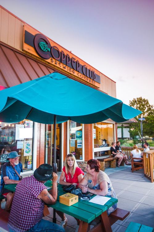 People enjoying drinks at Copper Club Brewery, seated under a green umbrella on a warm evening.