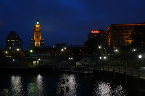 City skyline at night with illuminated buildings reflecting on water, under a cloudy sky.