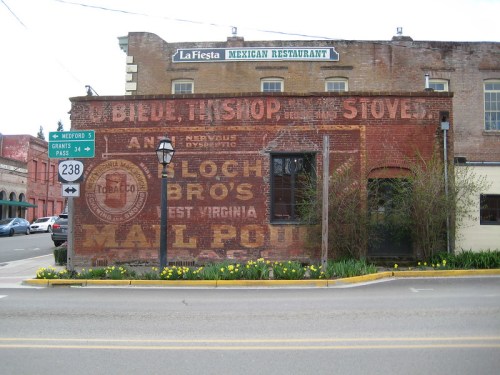 Faded vintage advertisements on a brick wall, with a street sign and blooming yellow flowers in the foreground.