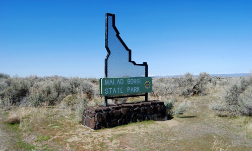 Sign for Malad Gorge State Park in Idaho, shaped like the state outline, surrounded by sagebrush and clear blue sky.