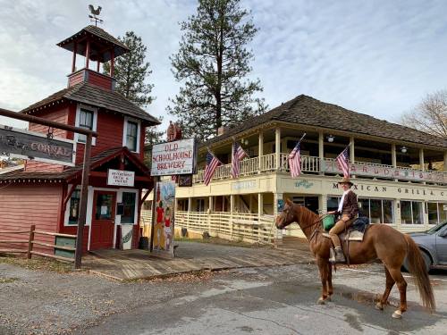 A person on horseback stands in front of a rustic building with American flags, near a brewery and restaurant.