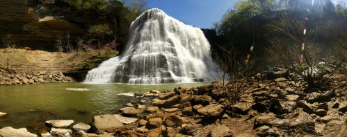 A panoramic view of a cascading waterfall surrounded by rocky terrain and greenery under a clear blue sky.