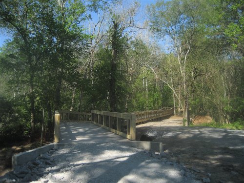 A wooden bridge spans a path through a lush green forest, surrounded by trees and clear blue skies.