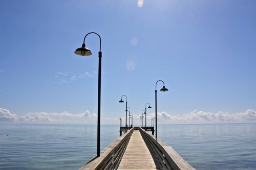 A wooden pier extends into calm waters under a clear blue sky, lined with street lamps.