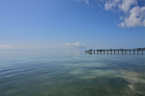 A calm, clear blue sea under a bright sky, with a wooden pier extending into the water.