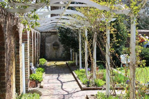 A serene garden path lined with greenery and a white trellis, leading to a stone wall and fountain.