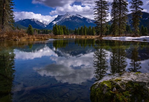 A serene mountain landscape reflected in a calm lake, surrounded by evergreen trees and a partly cloudy sky.