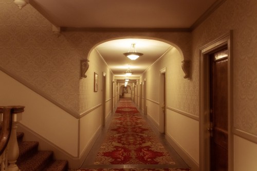 A dimly lit hotel hallway with patterned wallpaper, ornate light fixtures, and a red carpet leading to a distant door.