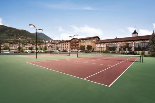 A tennis court with a green surface, surrounded by buildings and mountains under a clear blue sky.