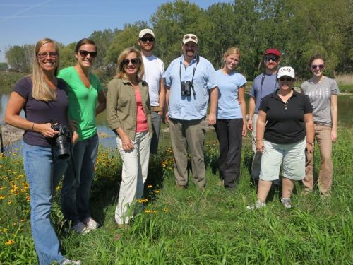 A group of eight people stands together outdoors near a pond, surrounded by greenery and wildflowers.