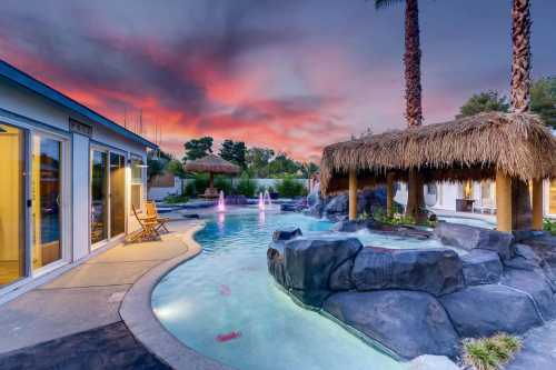 A serene pool area with a waterfall, palm trees, and a thatched hut, set against a colorful sunset sky.