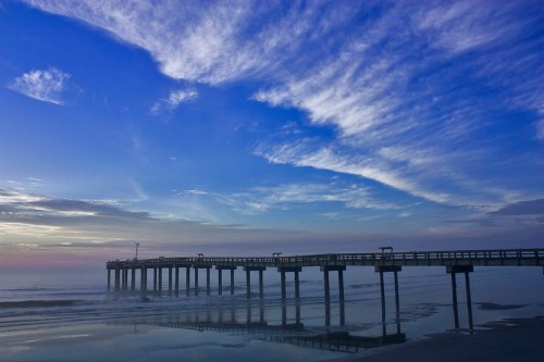 A serene pier extends into calm waters under a vibrant blue sky with wispy clouds at sunset.