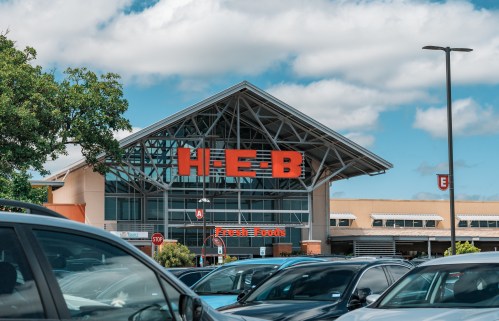 Exterior of an HEB grocery store with a clear blue sky and parked cars in the foreground.