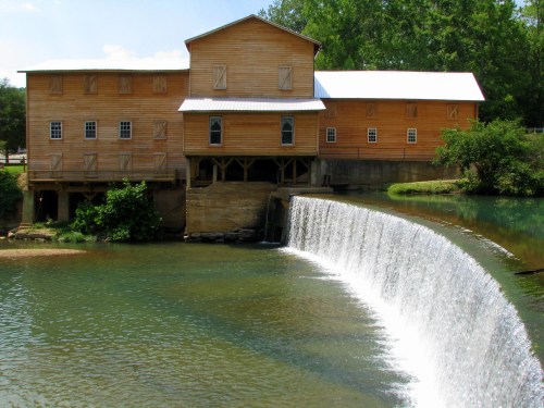 A wooden mill building beside a flowing waterfall, surrounded by greenery and a clear blue sky.