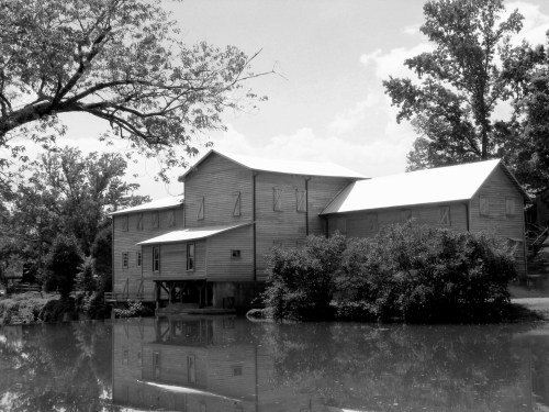 A historic wooden mill beside a calm pond, surrounded by trees, captured in black and white.