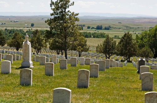 A serene cemetery with rows of white gravestones, surrounded by green grass and distant hills under a clear blue sky.