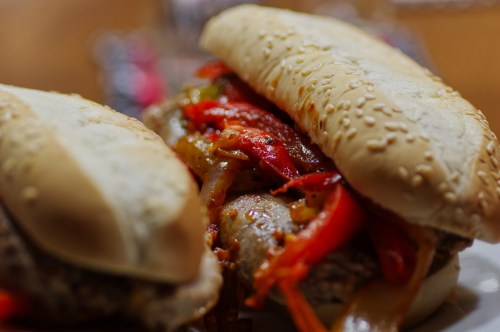 Two sesame seed buns filled with grilled sausage and colorful sautéed peppers, served on a plate.