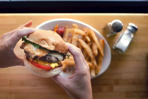 A person holds a burger with lettuce, tomato, and cheese, with a plate of fries in the background on a wooden table.