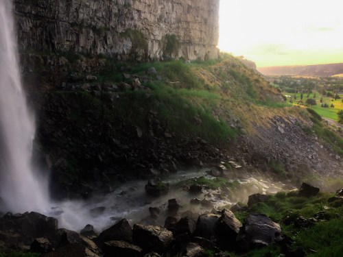 A waterfall cascades over rocky cliffs, surrounded by lush greenery and a misty river below, with a sunset in the background.