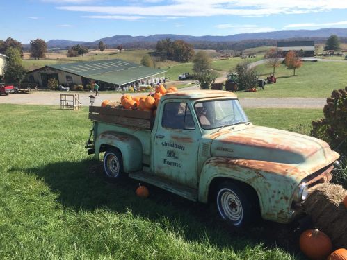 A vintage truck filled with pumpkins sits on a grassy field, with rolling hills and a farm in the background.