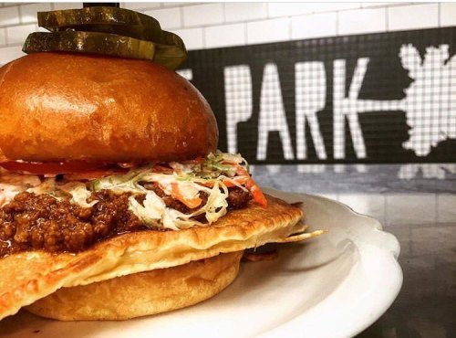 A close-up of a burger with pickles, lettuce, and tomato, set against a tiled background with the word "PARK."