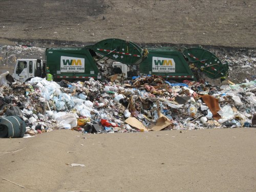Two waste management trucks parked near a large pile of garbage at a landfill site.