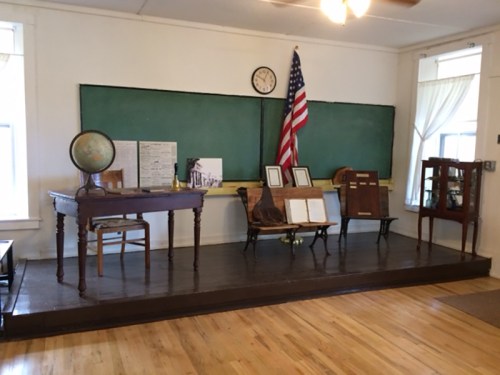 A vintage classroom setup featuring a wooden desk, globe, chairs, and an American flag against a green chalkboard.