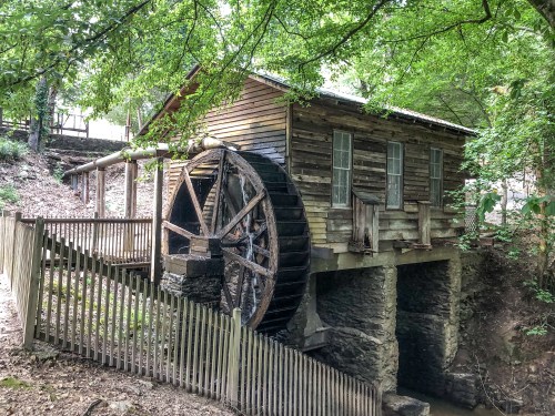 A rustic wooden watermill with a large wheel, surrounded by trees and a wooden fence.