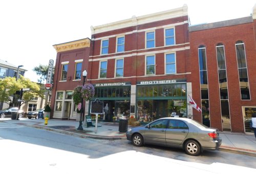 A brick building with "Harrison Brothers" storefront, featuring large windows and flower baskets, on a sunny street.