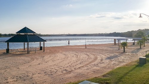 A sandy beach with a gazebo, a pier in the background, and calm water under a clear sky.