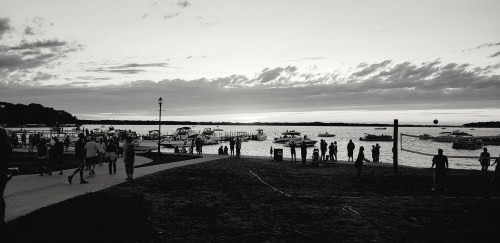 Silhouetted people walk along a waterfront at sunset, with boats on the water and clouds in the sky.