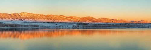 Sunset over snow-capped mountains reflecting in a calm lake, with a serene winter landscape.