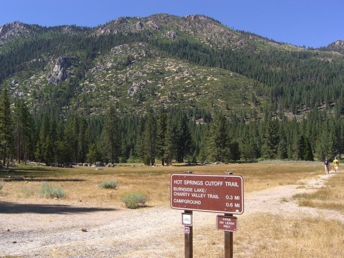 A trail sign for Hot Springs Cutoff Trail, with mountains and trees in the background, indicating nearby trails and campground.