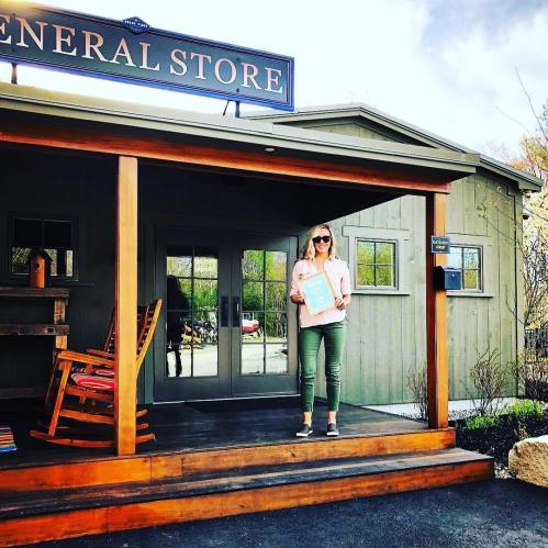 A woman stands on the porch of a general store, holding a sign, with a rocking chair nearby and trees in the background.