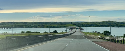 A wide road leads to a bridge over a river, surrounded by green hills and a cloudy sky.