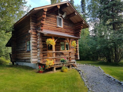 A charming log cabin surrounded by greenery, featuring flower baskets and a stone pathway leading to the entrance.